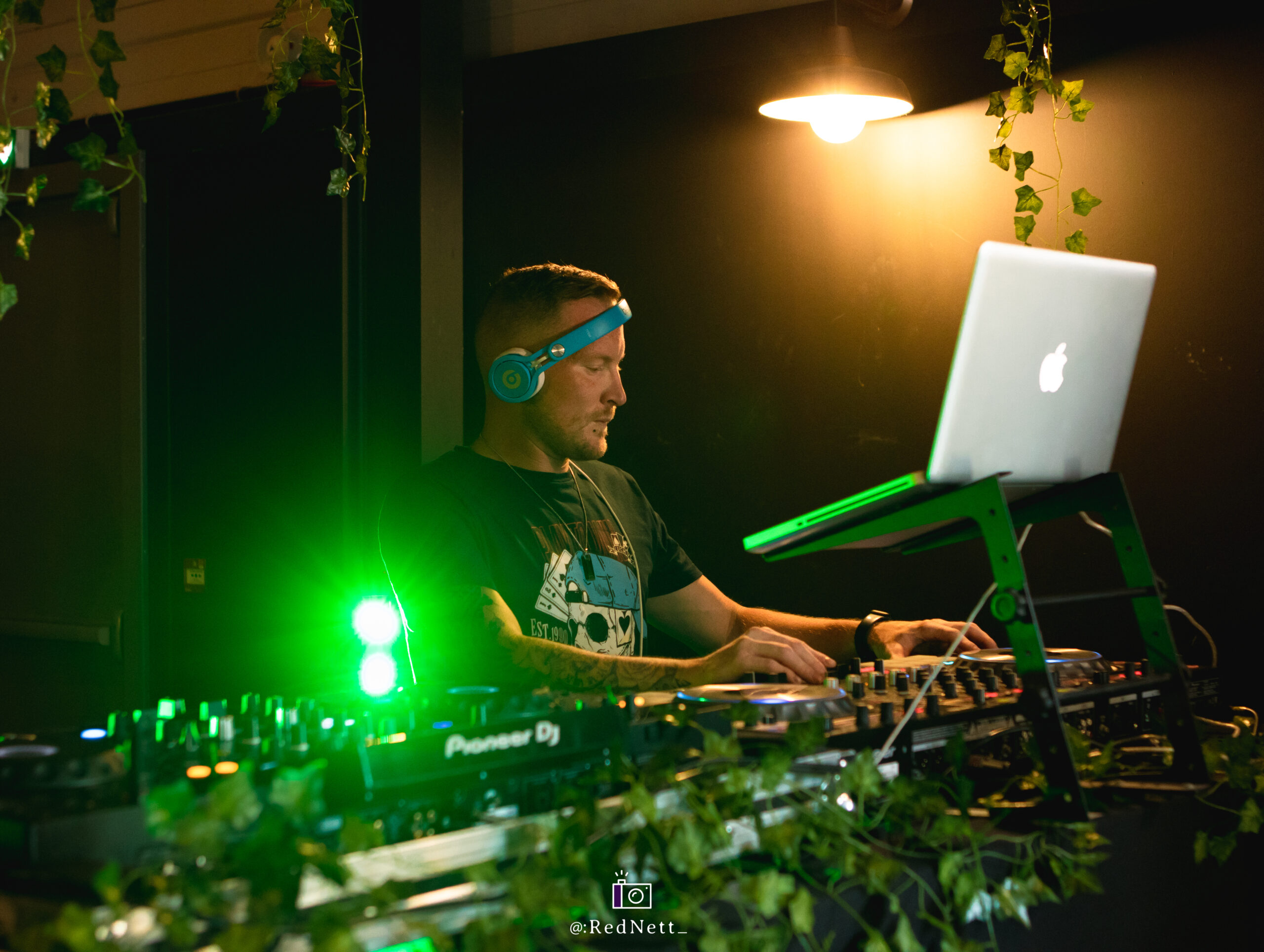 Photo d’un DJ en train de mixer dans un bar avec des plantes artificielles et un fond noir